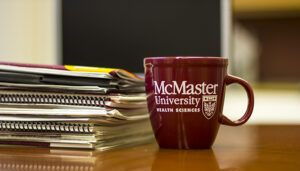 A McMaster coffee mug on a wooden desk next to a stack of notebooks